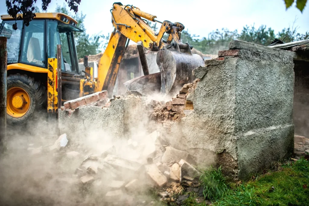 Excavator demolishing a concrete wall with dust rising from the debris